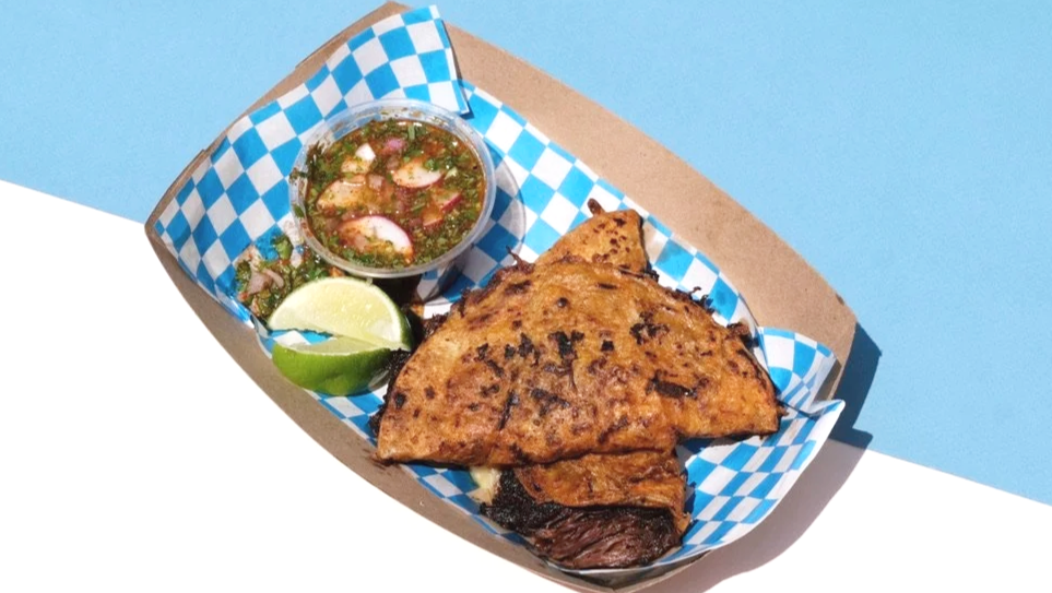 Shredded beef taco with side of dipping sauce all served on a checkered paper liner in a basket on a light blue and white background.