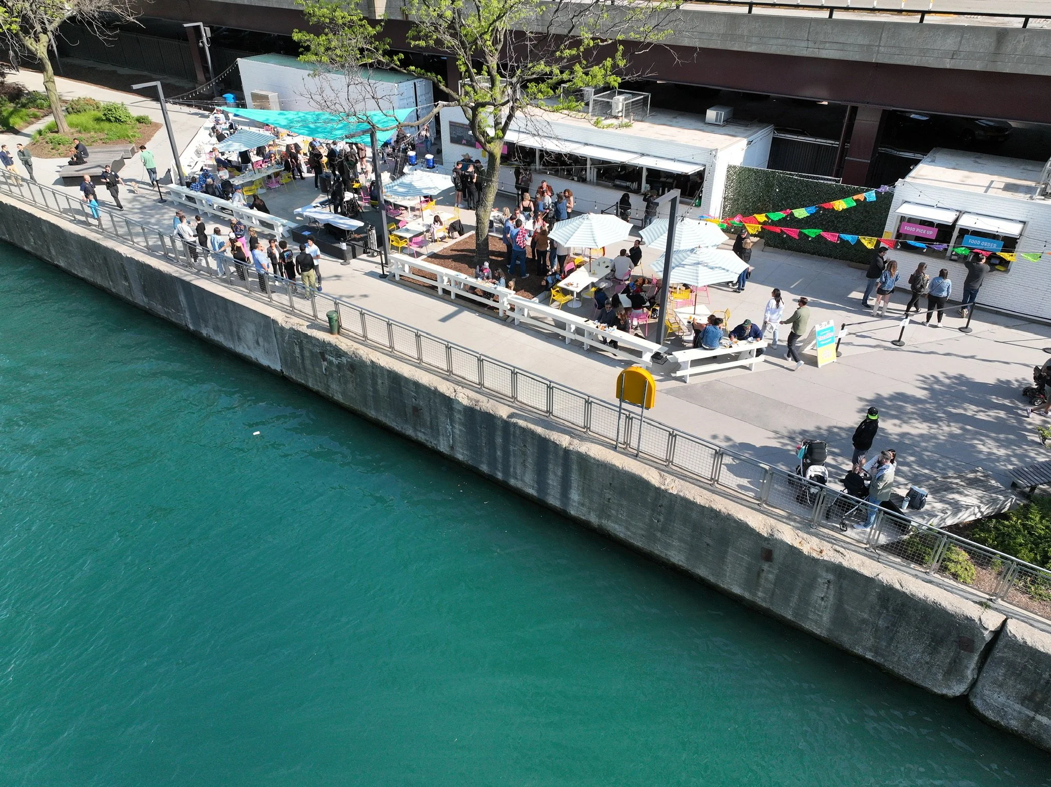 Aerial view of a riverwalk with a crowd gathered at outdoor food vendors and picnic tables, shaded by umbrellas, along a concrete waterfront with turquoise water in the foreground.