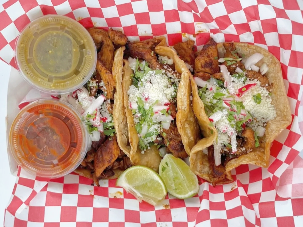A serving of Mexican street tacos on a red and white checkered paper with two lemon wedges, two small cups of sauce, and grilled meat and toppings including chopped onions and cilantro.