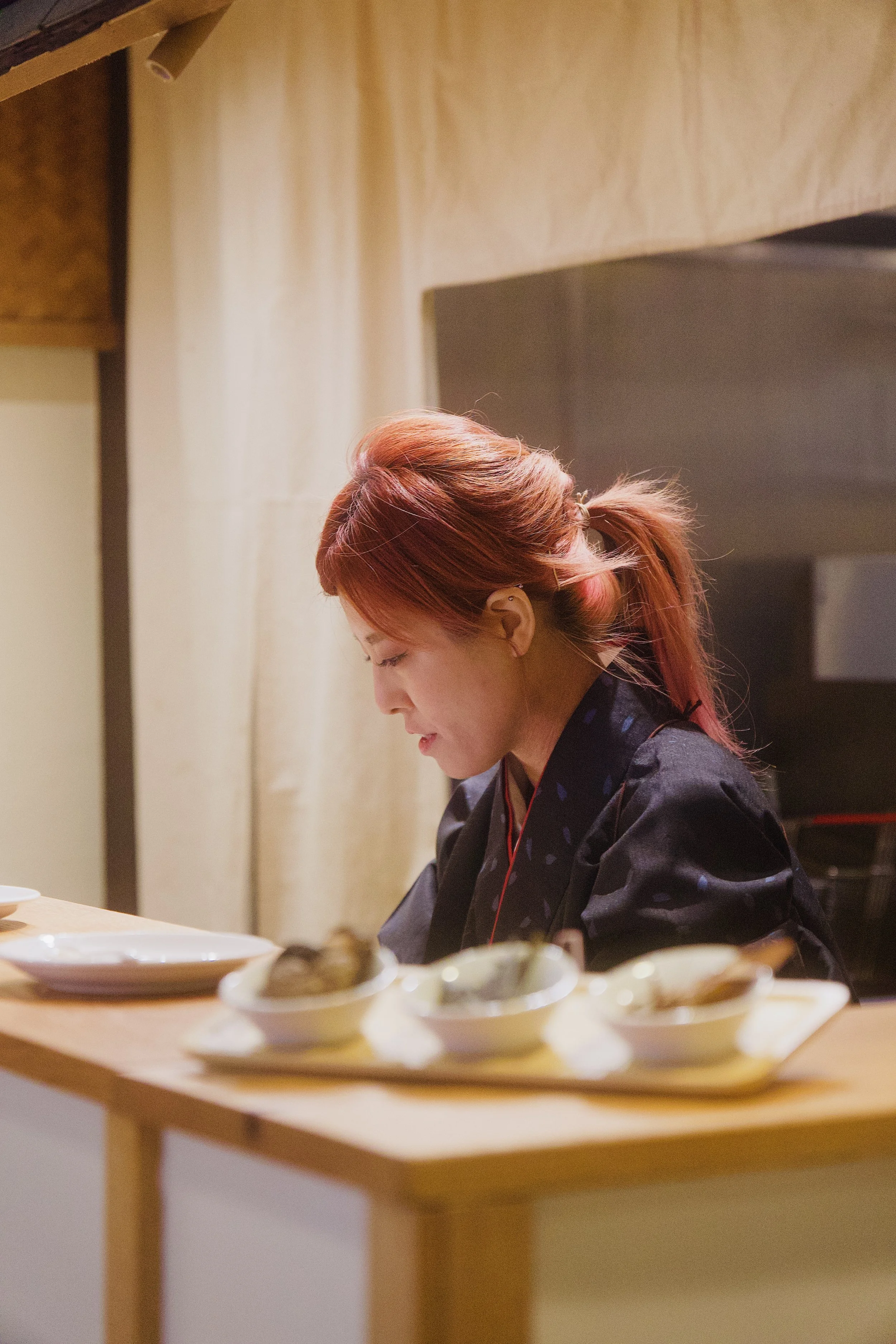 A woman with red hair tied in a ponytail, wearing a black traditional outfit, sitting at a wooden counter with small bowls of food in front of her.