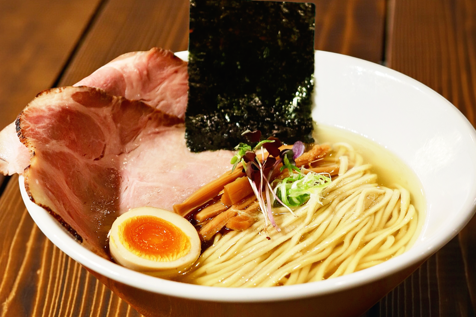 A bowl of ramen with slices of pork, half a soft-boiled egg, seaweed, bamboo shoots, green onions, and microgreens in broth, on a wooden table.