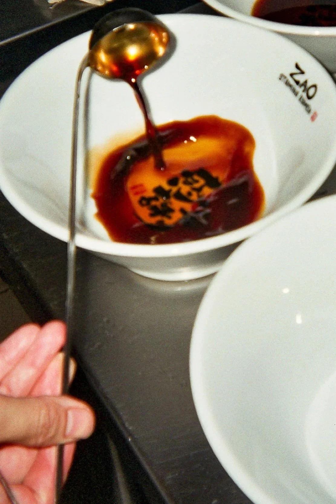 Pouring soy sauce from a spoon into a small white bowl with a ramen logo on the side.
