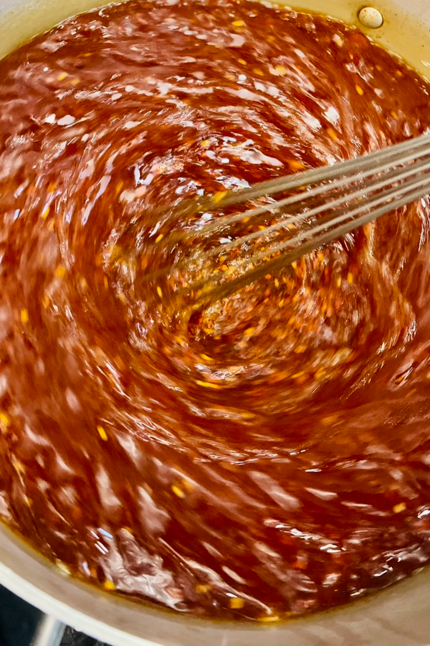 A close-up photo of a pot of spicy red chili sauce being stirred with a whisk.