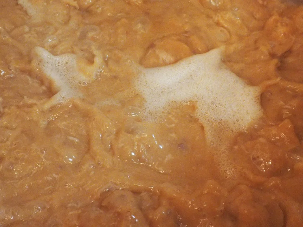 Close-up of boiling orange-colored liquid with foam, likely chicken cooking in a sauce or broth.