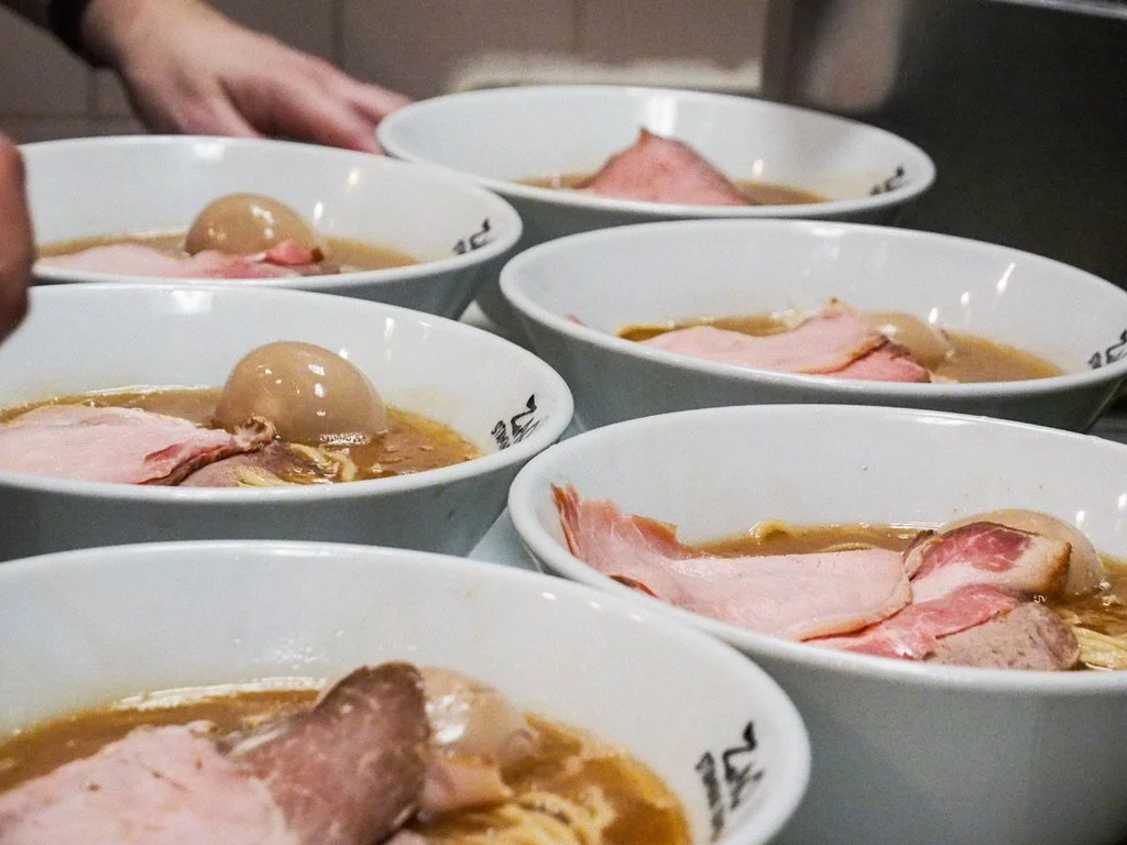 Six white bowls filled with ramen, topped with slices of pork, a soft-boiled egg, and broth, arranged on a table.