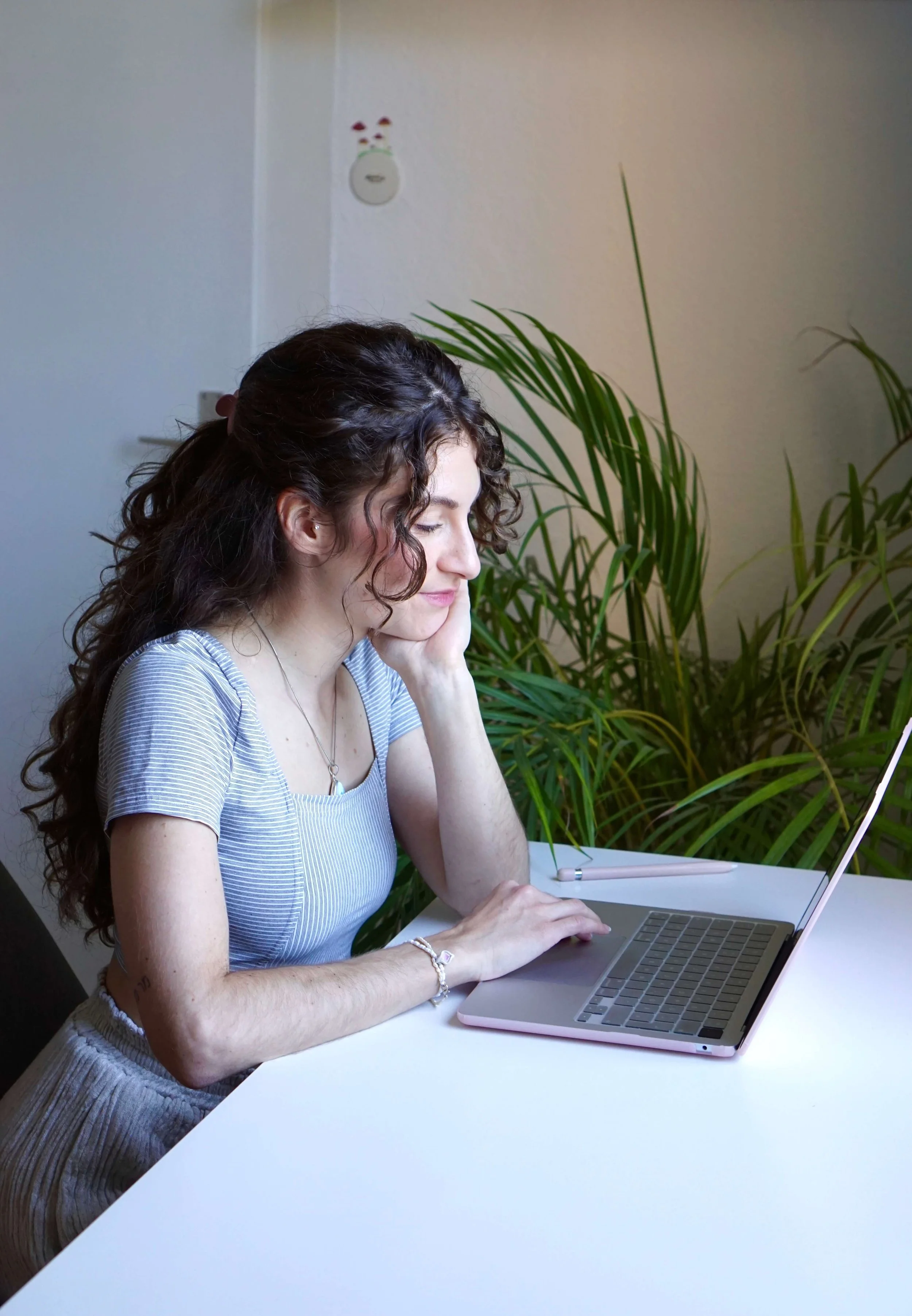 Sexualberaterin Mara Fink sitzt lächelnd während einer Online-Beratung am Laptop an einem weißen Tisch, während sie ihre Hand ans Kinn legt, im Hintergrund grüne Pflanzen und eine weiße Wand.