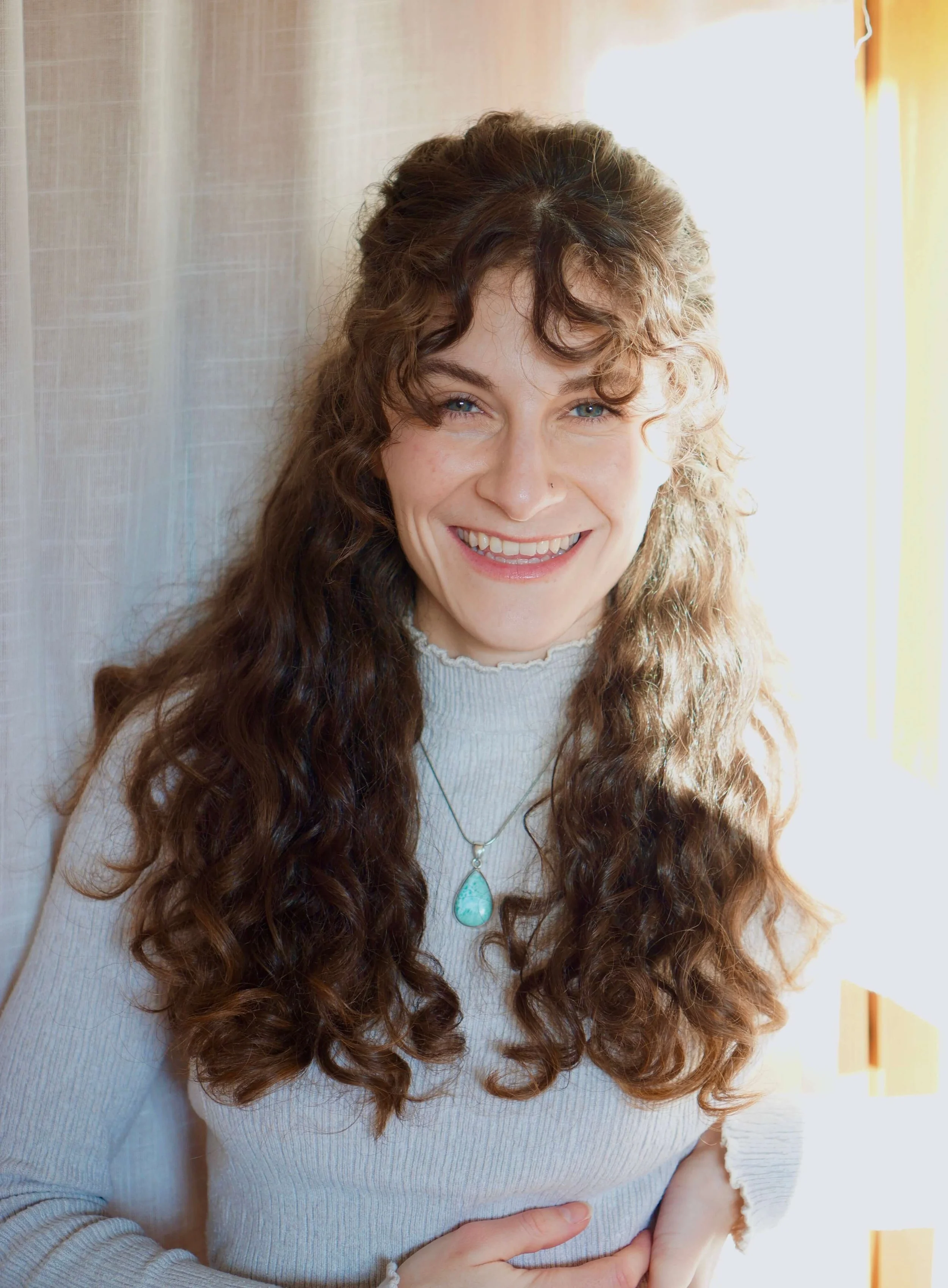 Sexual- und Paarberaterin Mara Fink mit lockigen braunen Haaren, die eine hellblaue Halskette trägt steht lächelnd vor einem hellen Hintergrund.