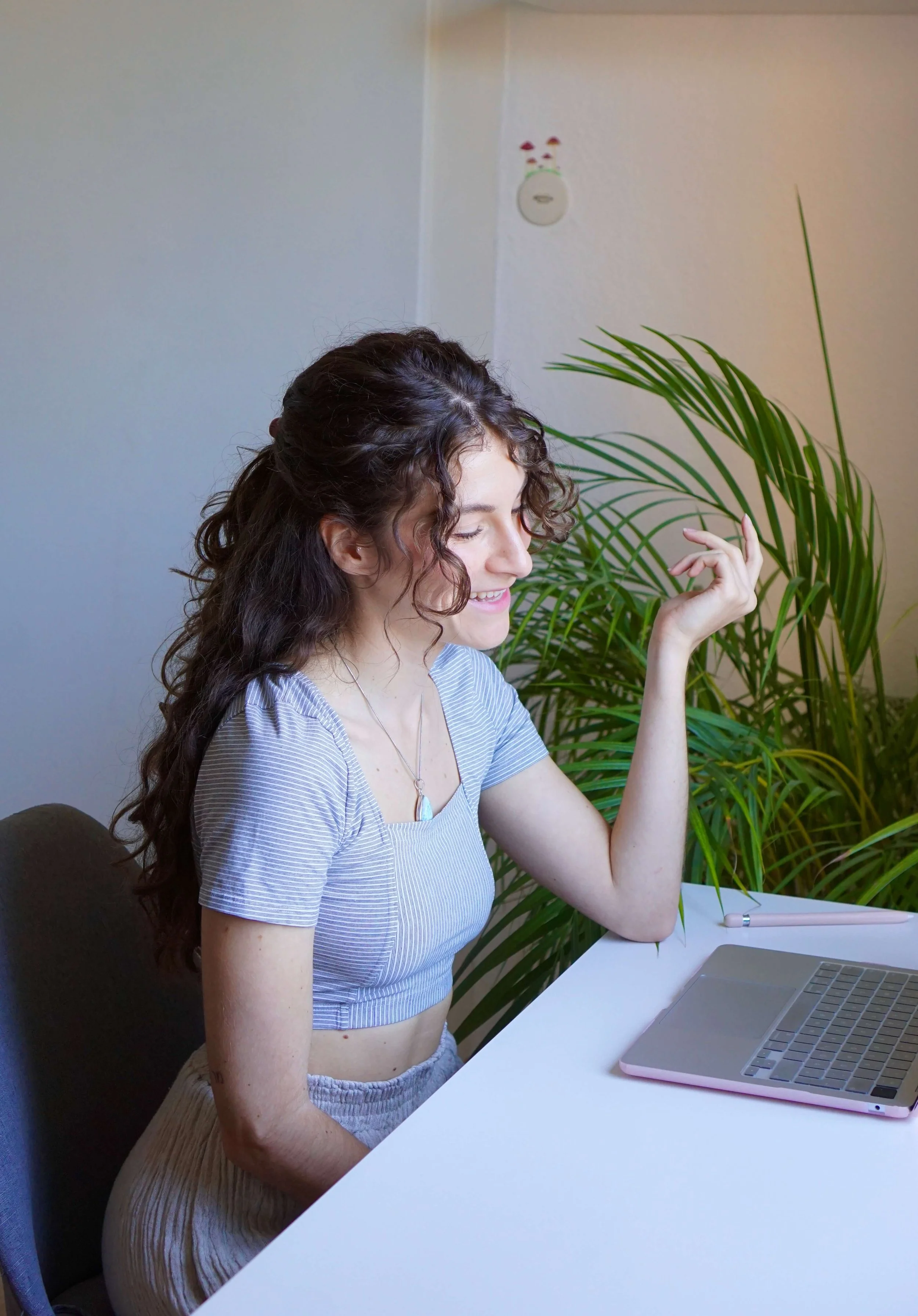 Sexualberaterin und Paarberaterin Mara Fink von spaceforyou mit Locken sitzt an einem weißen Tisch, führt auf einem Laptop eine Online Beratung durch, trägt ein gestreiftes Crop-Top und lächelt während sie die Hand hebt.