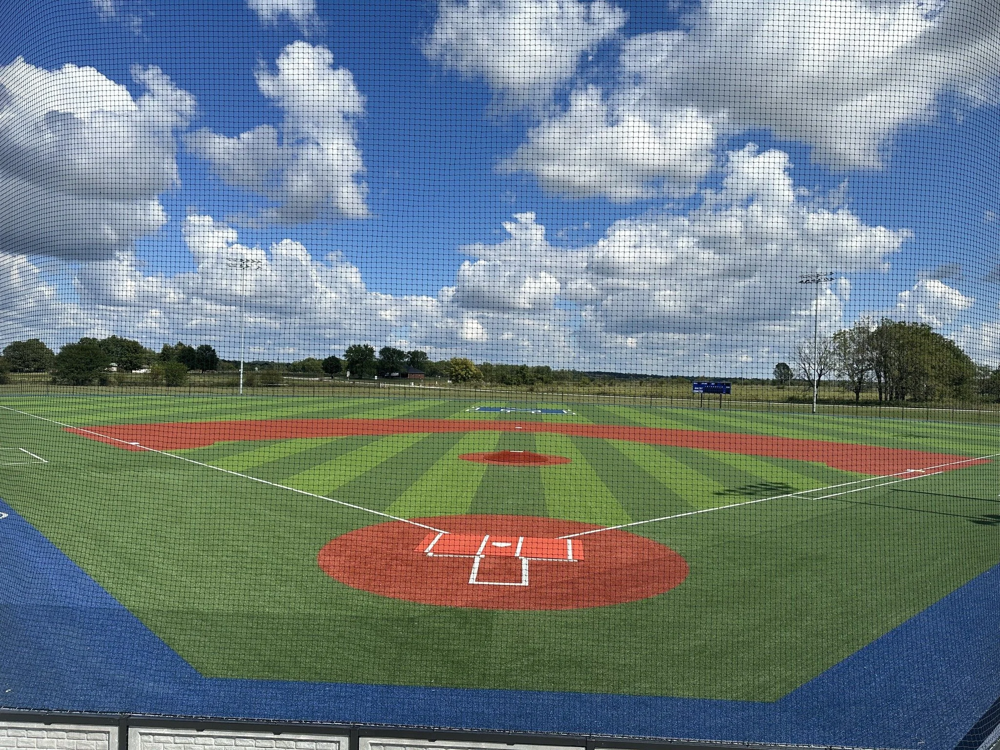 Empty baseball field viewed through a protective screen, with a blue sky filled with white clouds and trees in the background.