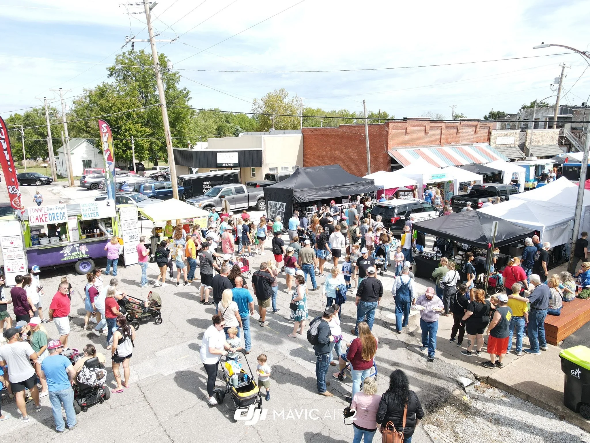 Outdoor market with people walking among tents and food trucks in a parking lot, green trees in the background, and a clear sky.