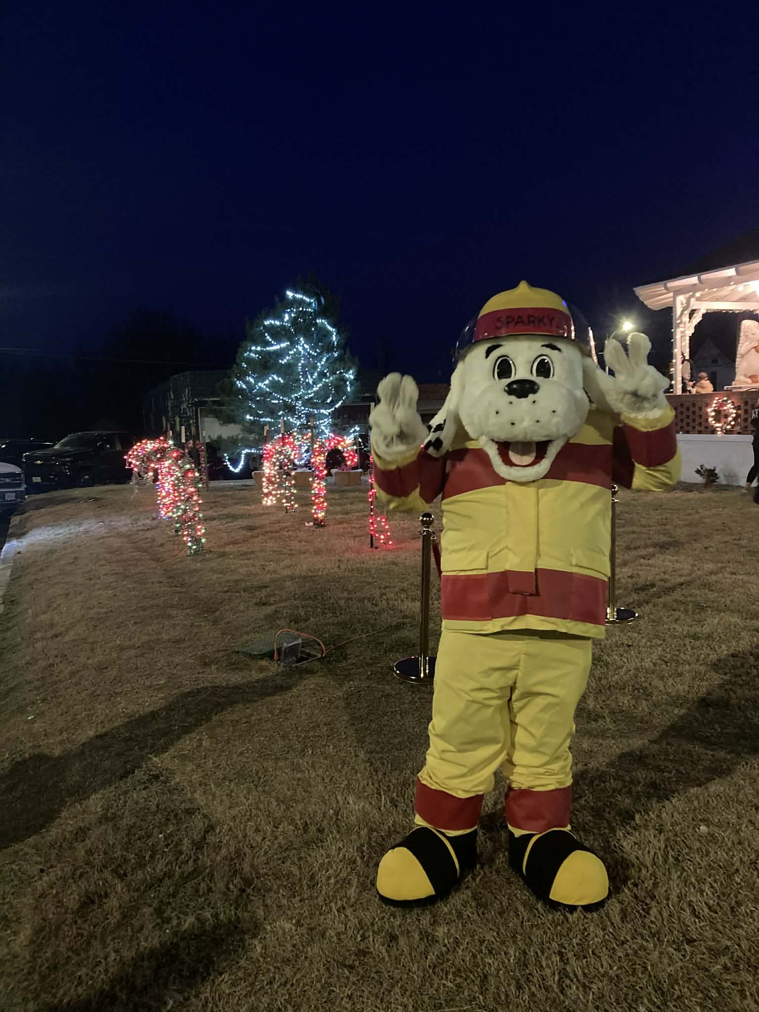 A person dressed in a firefighter-themed dog costume with a hat, yellow jacket, and pants, standing outdoors at night with Christmas decorations, a lit Christmas tree, and candy cane-shaped lights in the background.