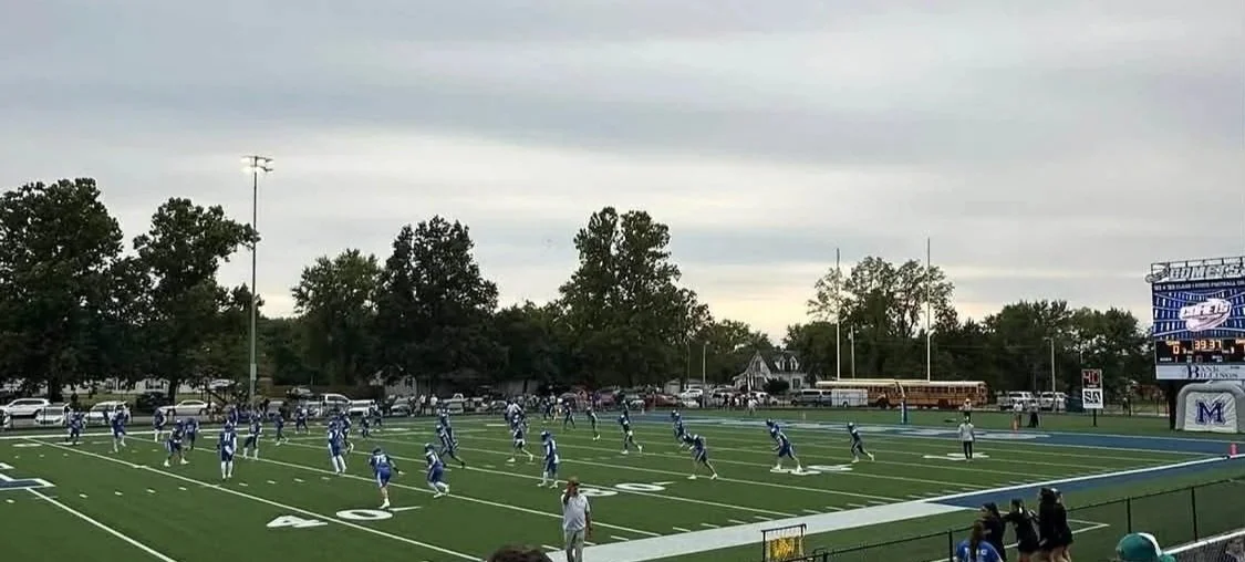 A football game in progress on a field with players in blue and white uniforms. The teams are lined up, preparing for play. Spectators and parked cars are visible in the background, along with a scoreboard showing the score and time. Trees line the perimeter of the field.