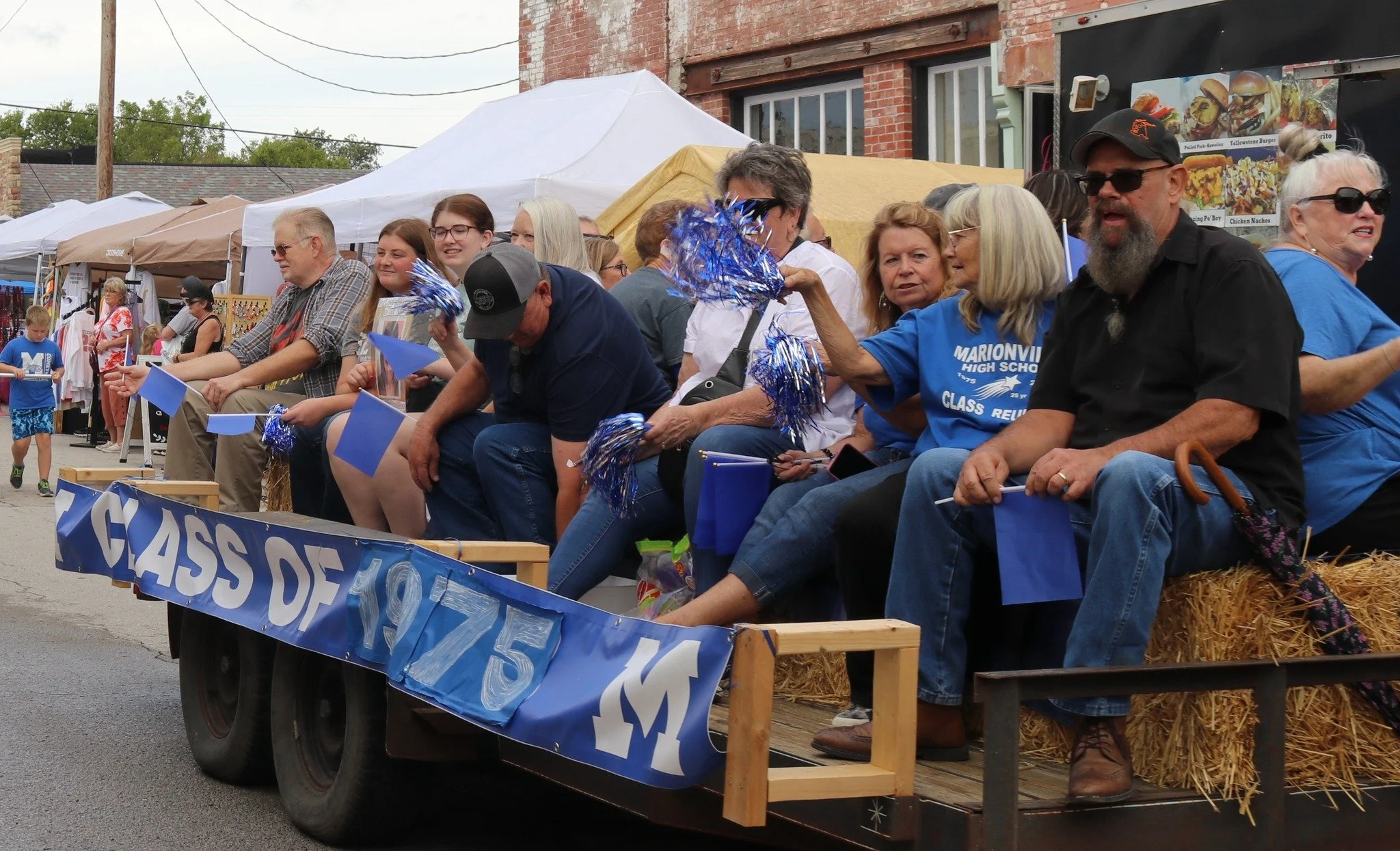 A parade float with the words "Class of 1975" is decorated with blue banners, small flags, and blue tinsel, carrying a group of people celebrating a class reunion in front of a street lined with tents and storefronts.