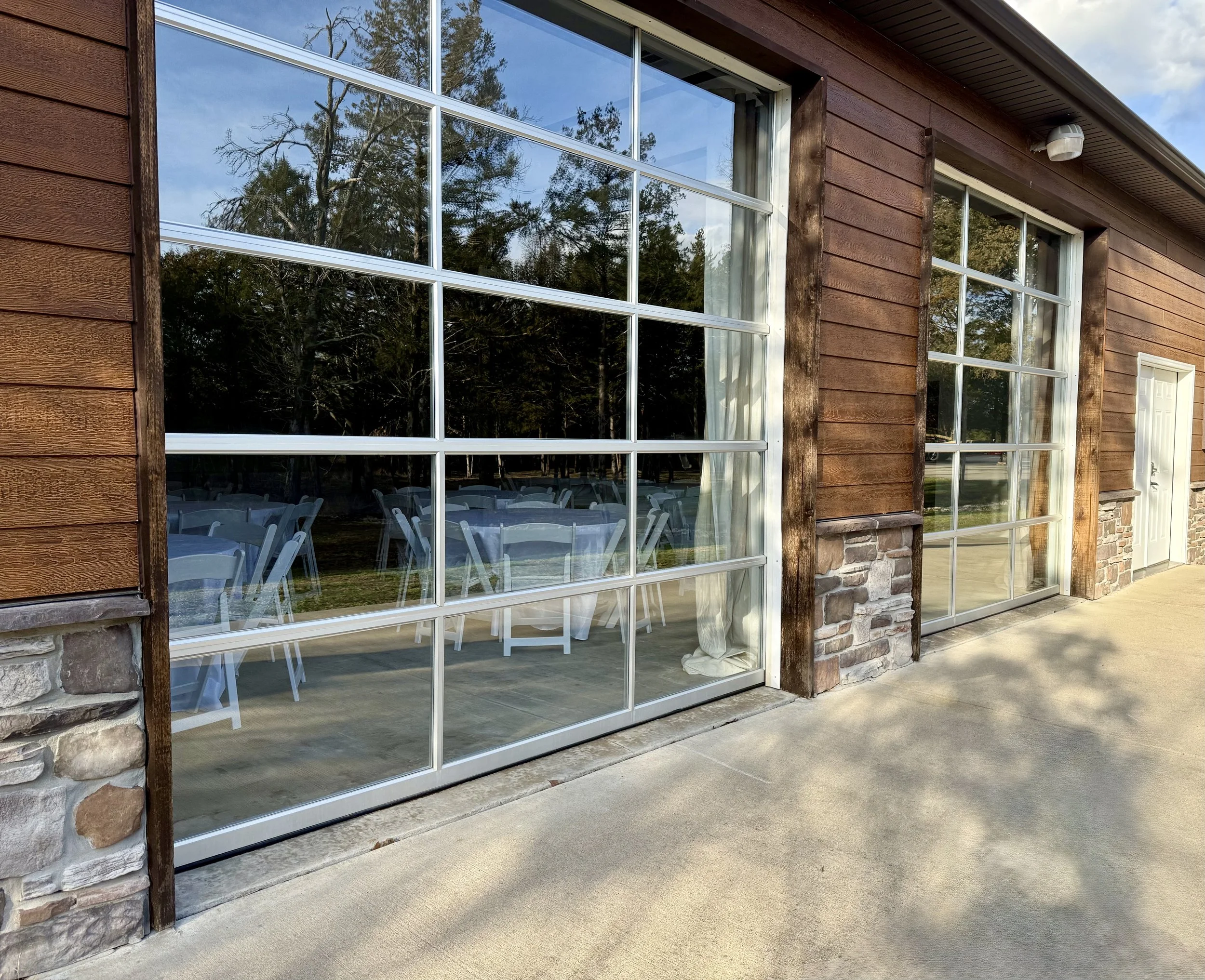Large window of a building with wooden and stone exterior, showing reflection of outdoor trees and tables inside with white chairs and tablecloths, on a concrete patio.