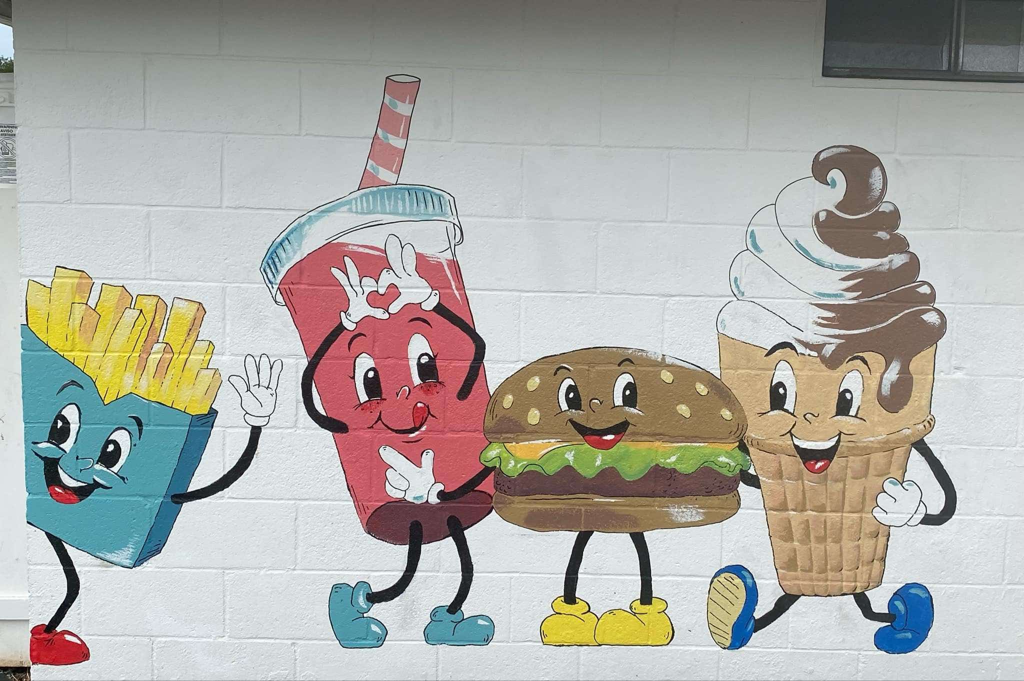 Colorful mural of animated food characters on a white brick wall. From left to right: a blue container of French fries with a smiling face, a red drink cup with a straw and a smiling face giving a peace sign, a cartoon hamburger with a smiling face, 
