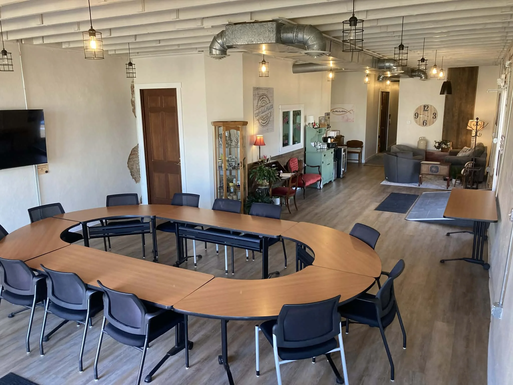 Spacious room with wooden flooring and ceiling, includes a large U-shaped conference table with black chairs, a wall-mounted TV, a sitting area with couches and armchairs, a clock, and various decorative items and lamps.