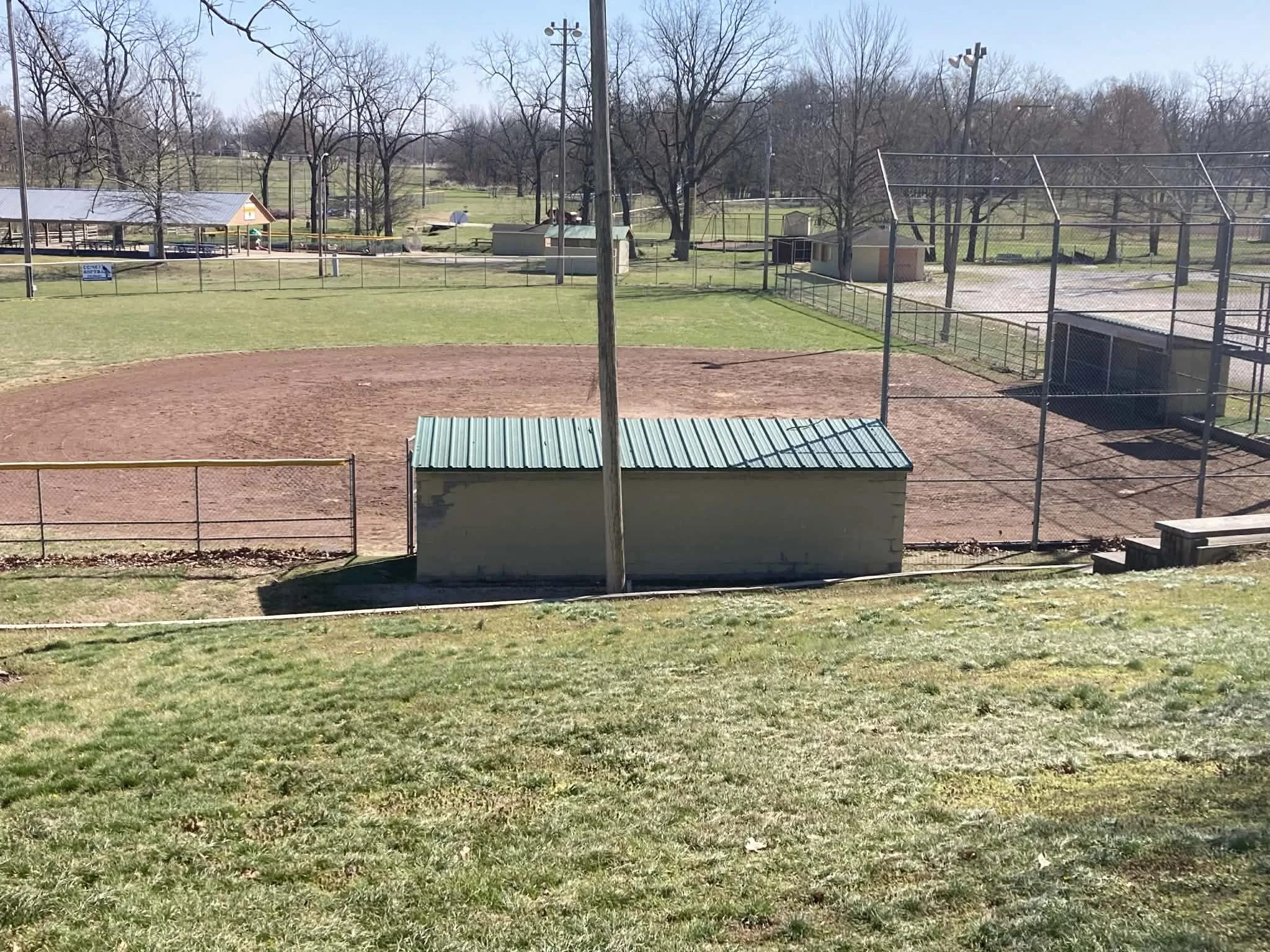 Empty baseball field with a dirt infield surrounded by a chain-link fence, a small building at the edge, and trees in the background. Marionville, MO City Park