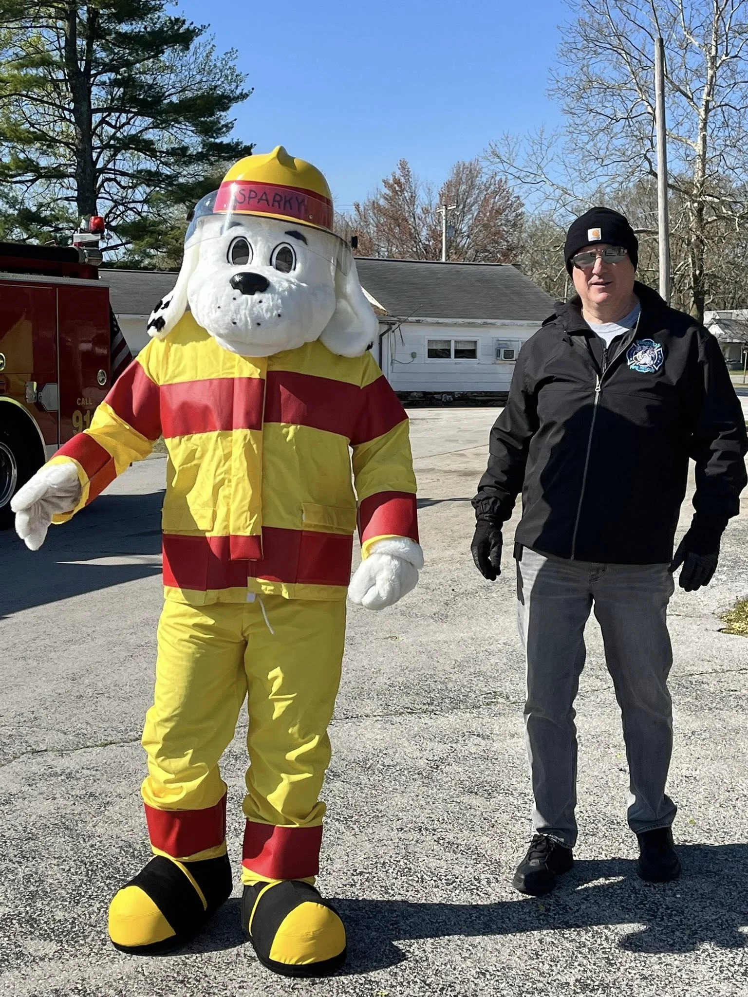 A person in a black jacket and gray pants standing next to a large costumed dog character dressed in firefighter gear, with a fire truck in the background.