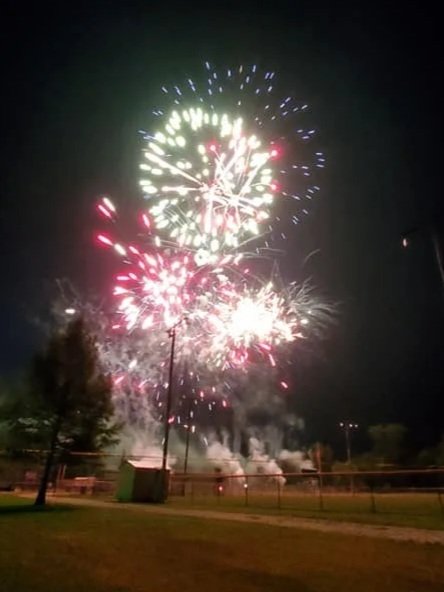 Fireworks exploding in the night sky above a grassy area with trees, a fence, and utility poles.