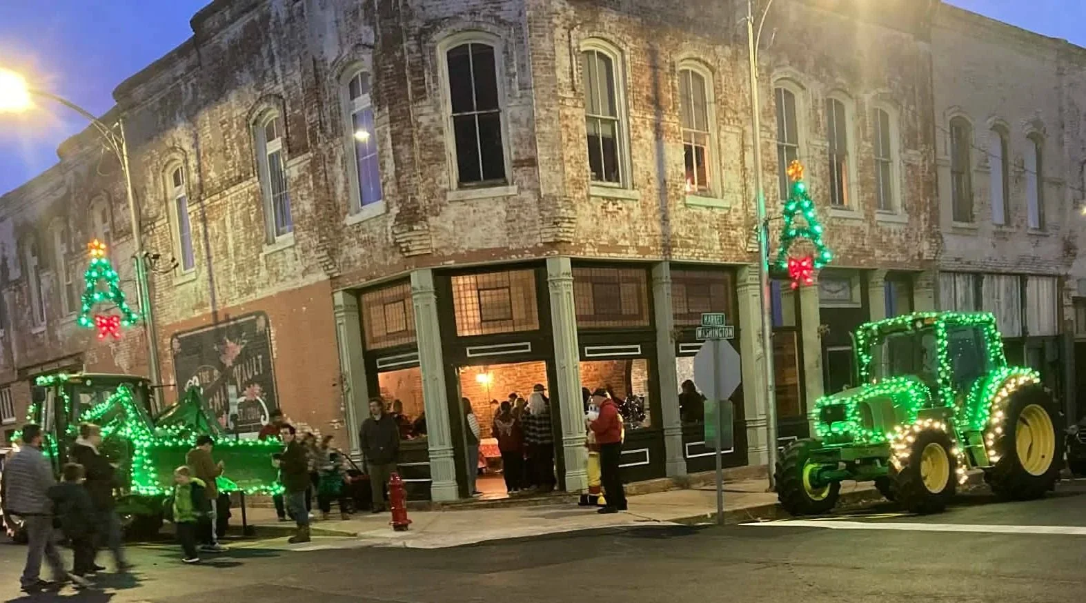 Street view of a corner building decorated with Christmas lights, including illuminated green tractors with lights, and Christmas tree-shaped lights on lampposts. People are gathered outside, some entering a store with large front windows, and the sc
