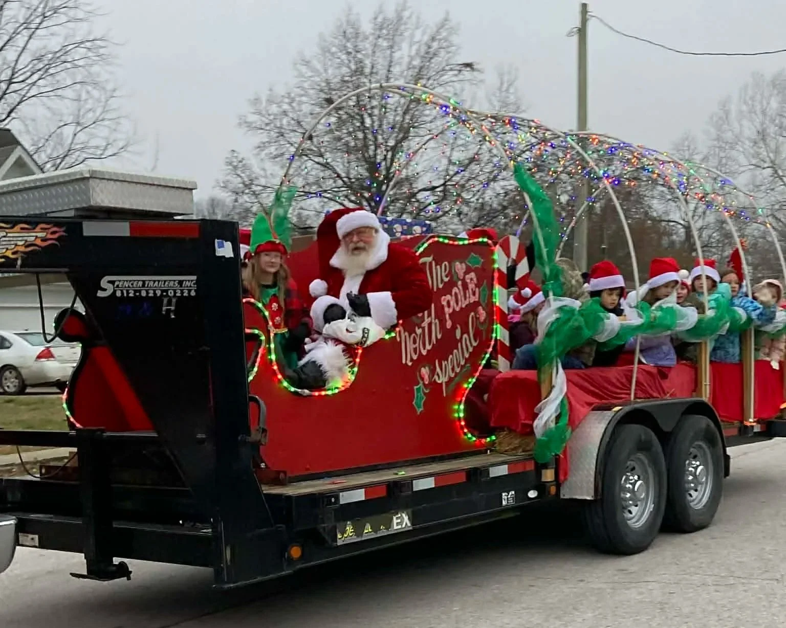 A Christmas parade float with Santa Claus, children in Santa hats, and festive decorations, being pulled by a black trailer.
