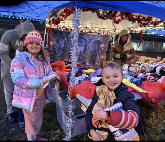 Two children smiling at a Christmas market stall decorated with tinsel and Christmas lights, with a vendor in the background.