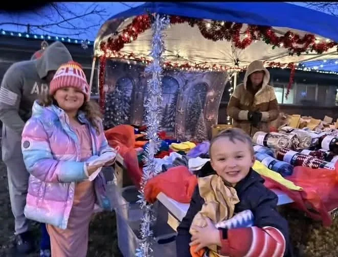 Two children smiling at an outdoor holiday market, standing in front of decorated stall with holiday lights, tinsel, and various items for sale, during evening.