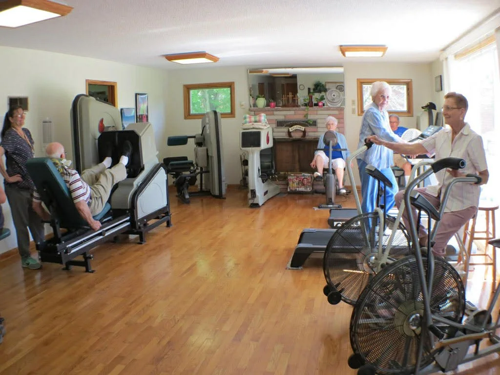 A room in a senior care facility with elderly people on exercise equipment and a staff member assisting.