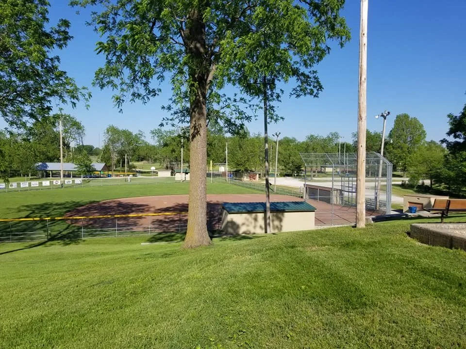 A baseball field with a brown dirt infield, green grass, and tall trees surrounding the area in a park. There are light poles, a wooden bench, and a small school building visible in the background.