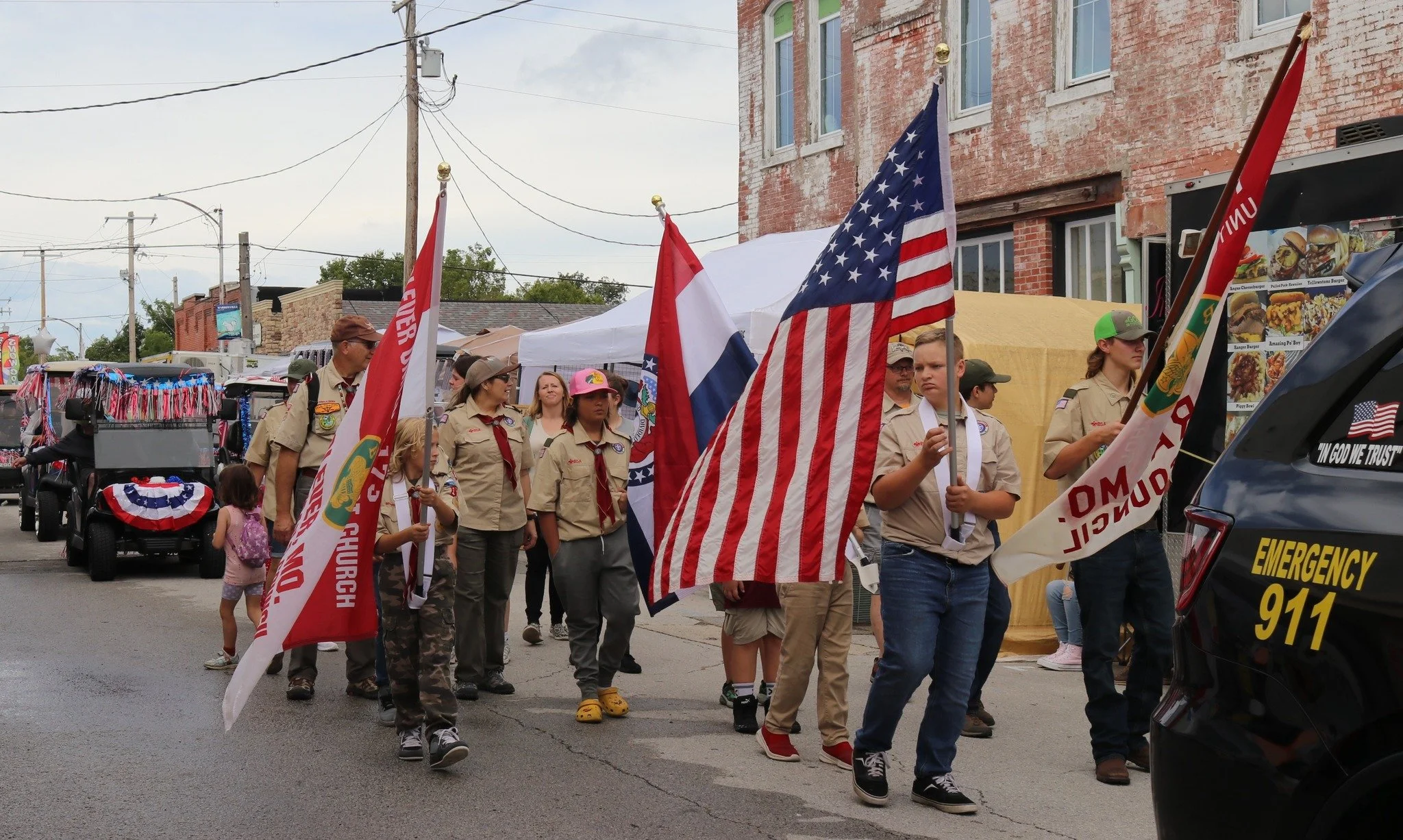 Group of people, including children and adults, participating in a parade or procession, carrying flags such as the American flag and Boy Scout flags, walking along a street with tents and buildings in the background.