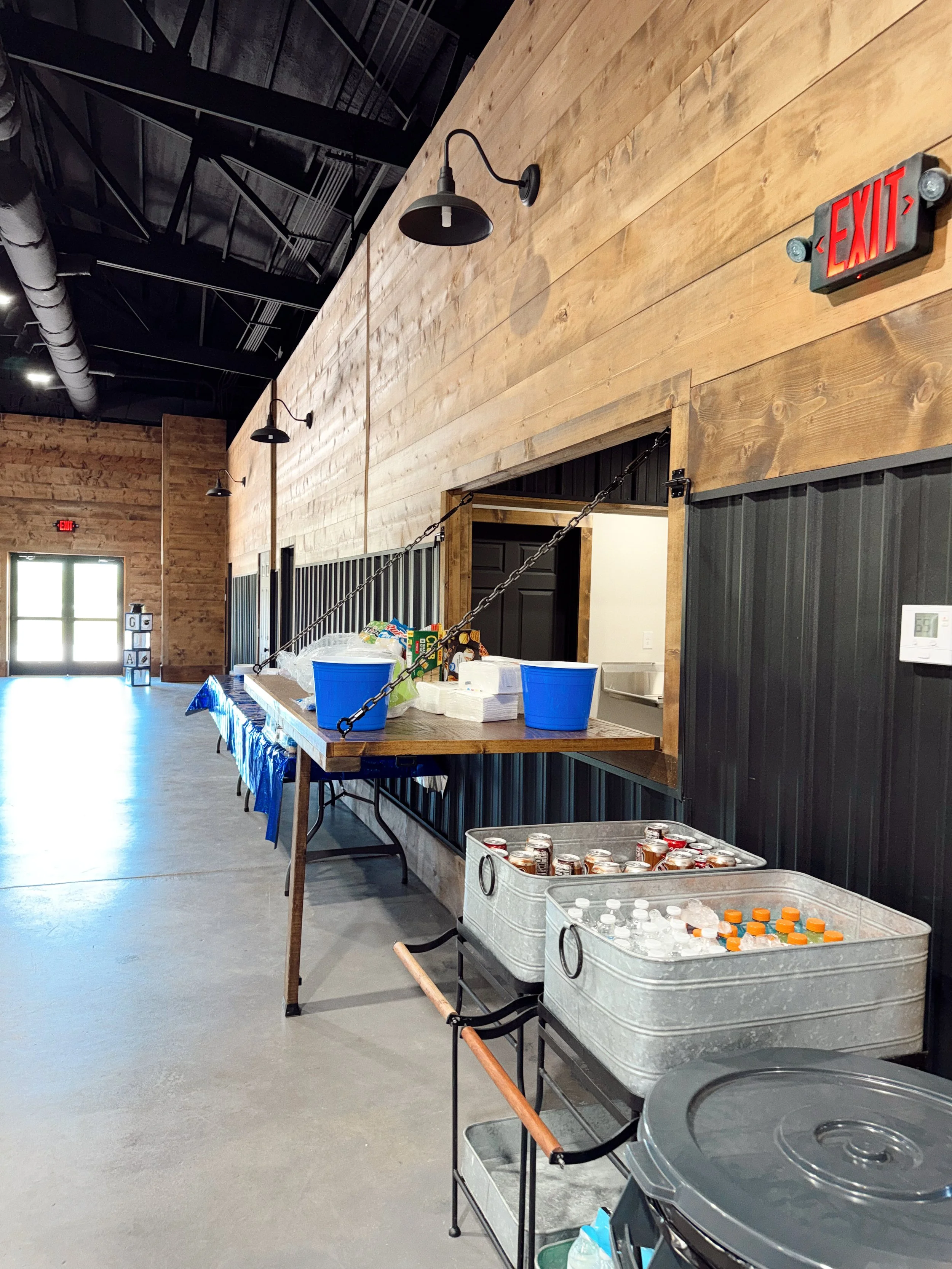 Indoor space with wooden walls and black ceiling, set up for a gathering or event, featuring food and drinks on tables, with some canned beverages in metal tubs, and a chain barrier in the foreground.