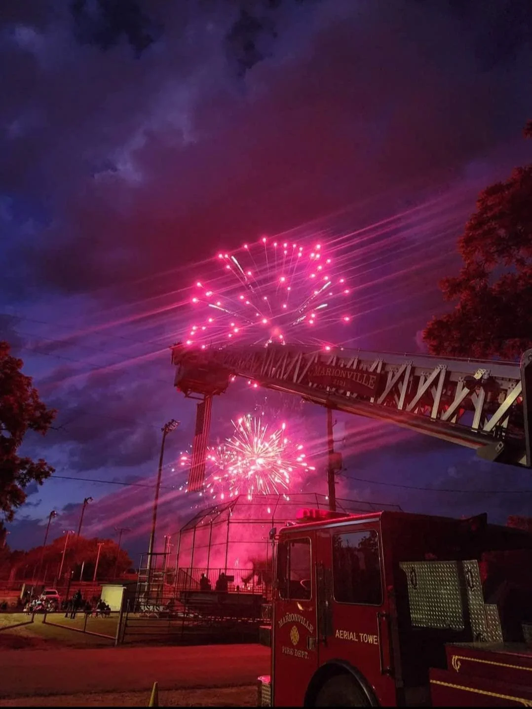 Fireworks display at night with a fire truck and a ladder in front, colorful explosions lighting up the dark sky.