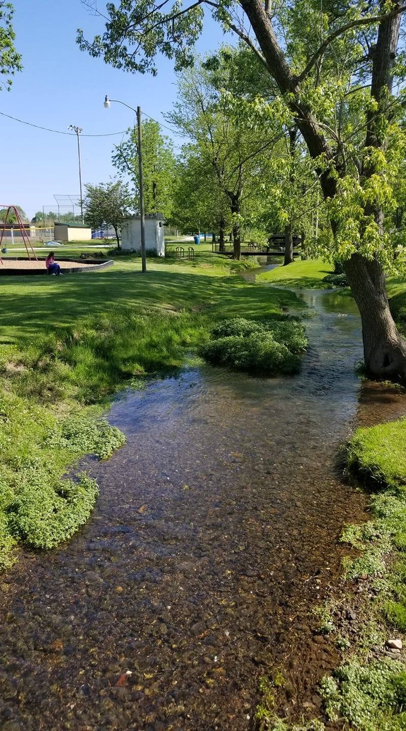 A park scene with a small creek, trees, a playground with swings, and a baseball field in the background on a sunny day.