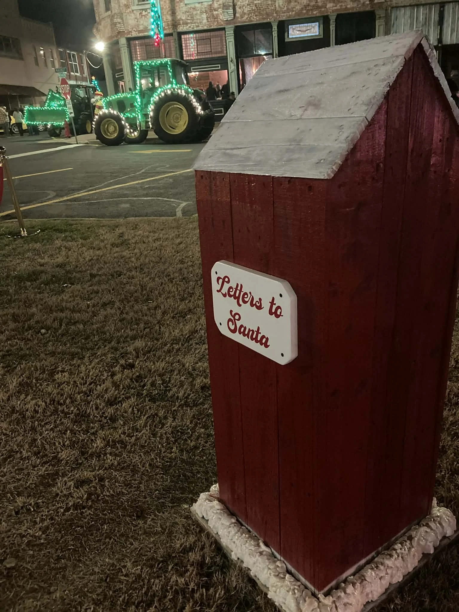 A red wooden mailbox with a white sign that reads "Letters to Santa" on a grassy area at night, with a parade float in the background featuring a tractor decorated with green lights.