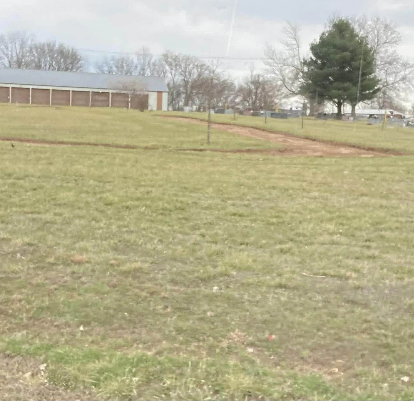 A grassy field with a dirt pathway leading up a small hill, a building to the left, tall trees in the background, and overcast sky.
