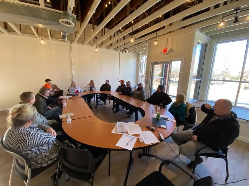 People seated around a large oval conference table in a well-lit room with windows, engaging in a meeting or discussion.