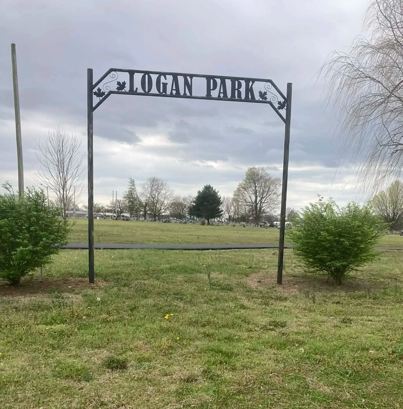 The entrance sign of Logan Park with an archway and metal lettering, flanked by green bushes, under a cloudy sky with mostly leafless trees in the background.