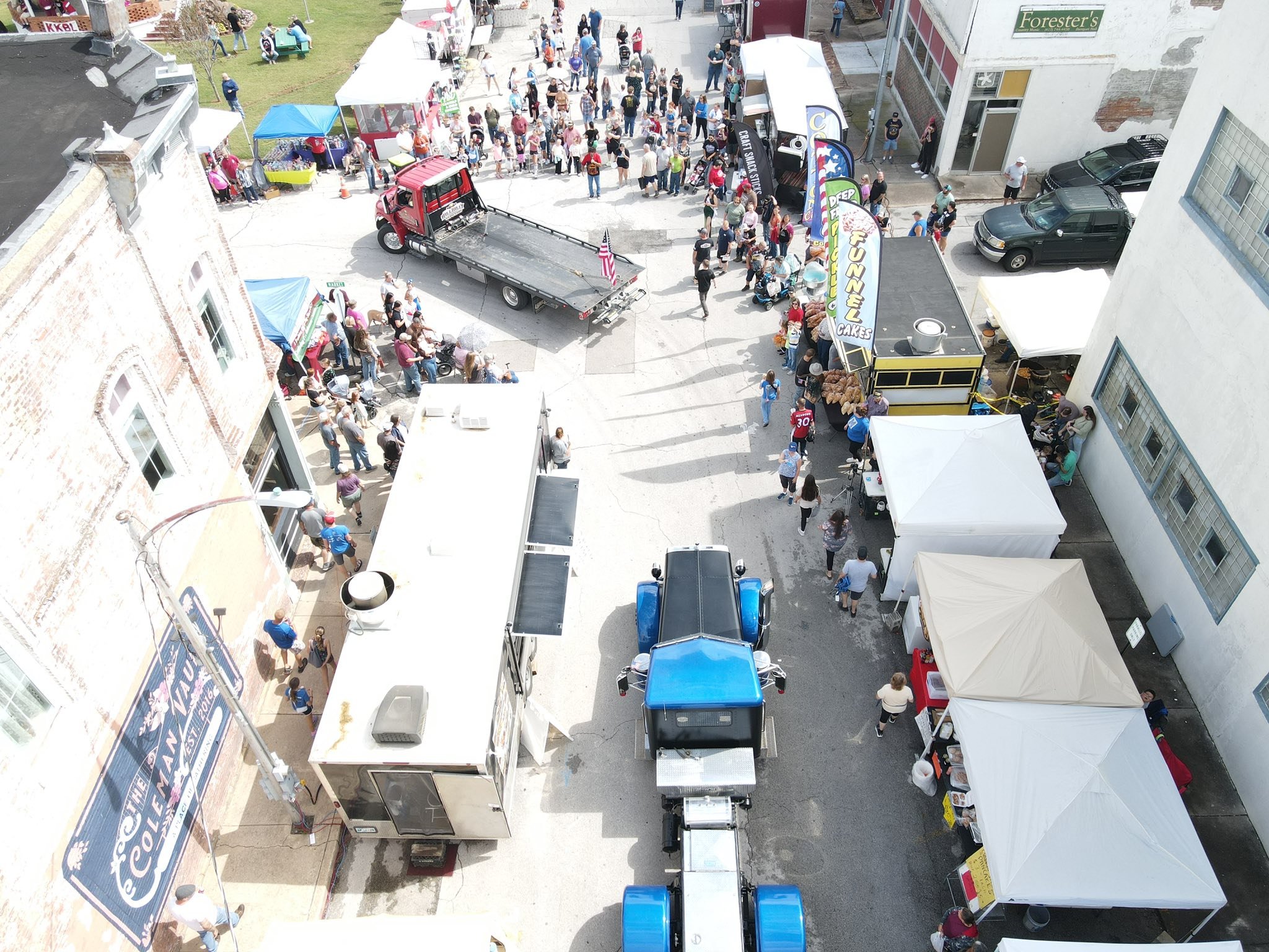 Overhead view of a street fair with food trucks, tents, and booths. People are walking and browsing, with a large crowd near the food vendors and a stage with horseshoe and carnival rides in the background.