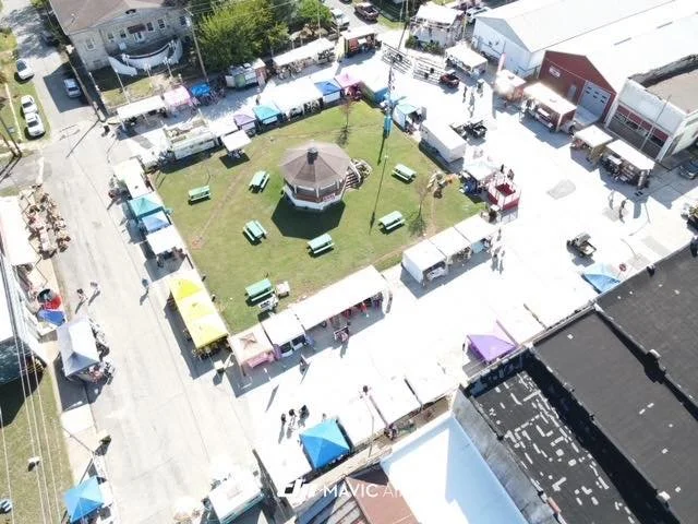 An aerial view of a small outdoor fair or market with a circular green space in the center, surrounded by vendor tents, tables, and food trucks.