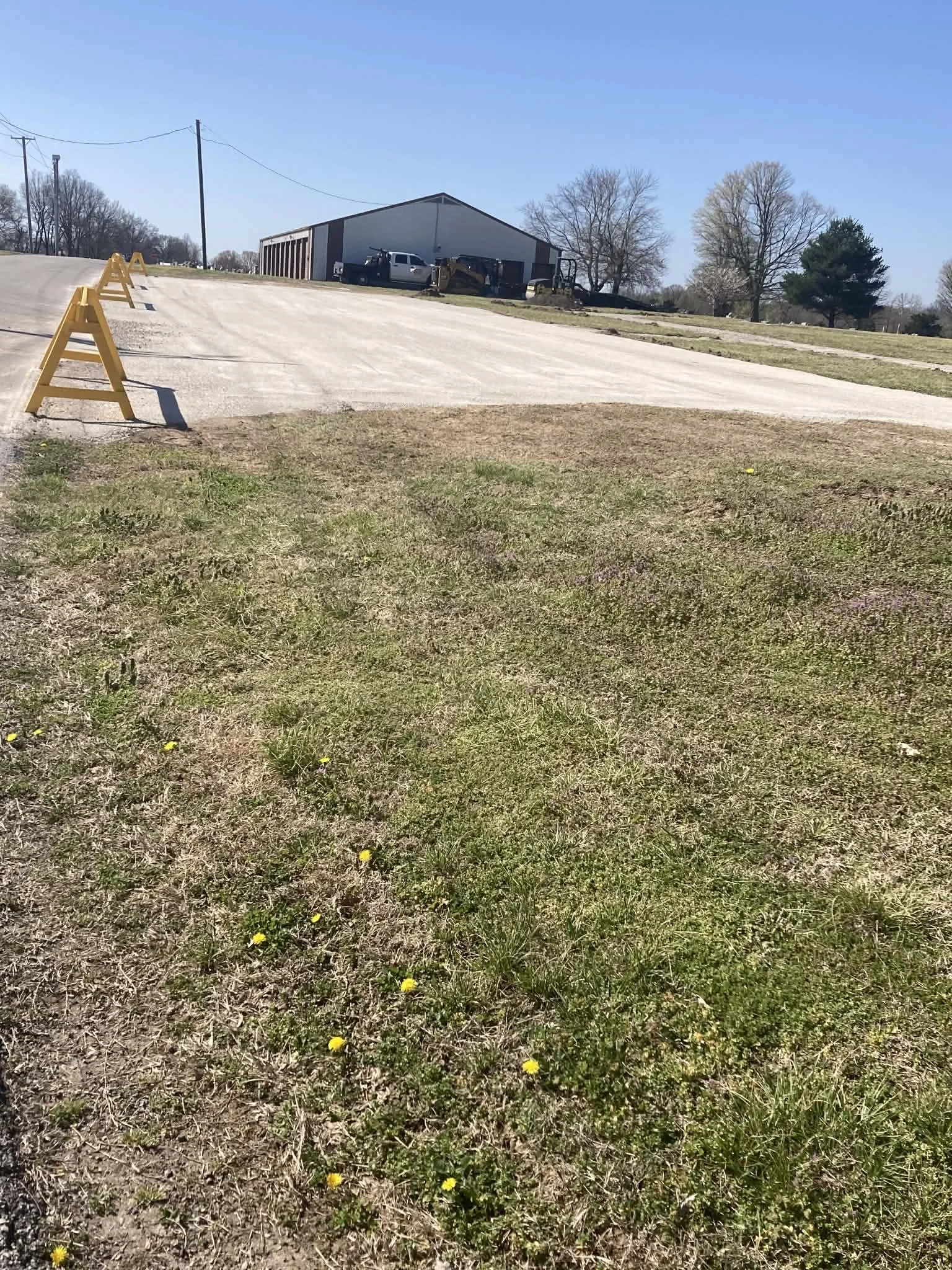 A sloped parking lot surface with yellow barriers along the edge. In the background, there is a large warehouse building, construction equipment, and parked vehicles. Surrounding the area are leafless trees and a grassy foreground with small yellow f