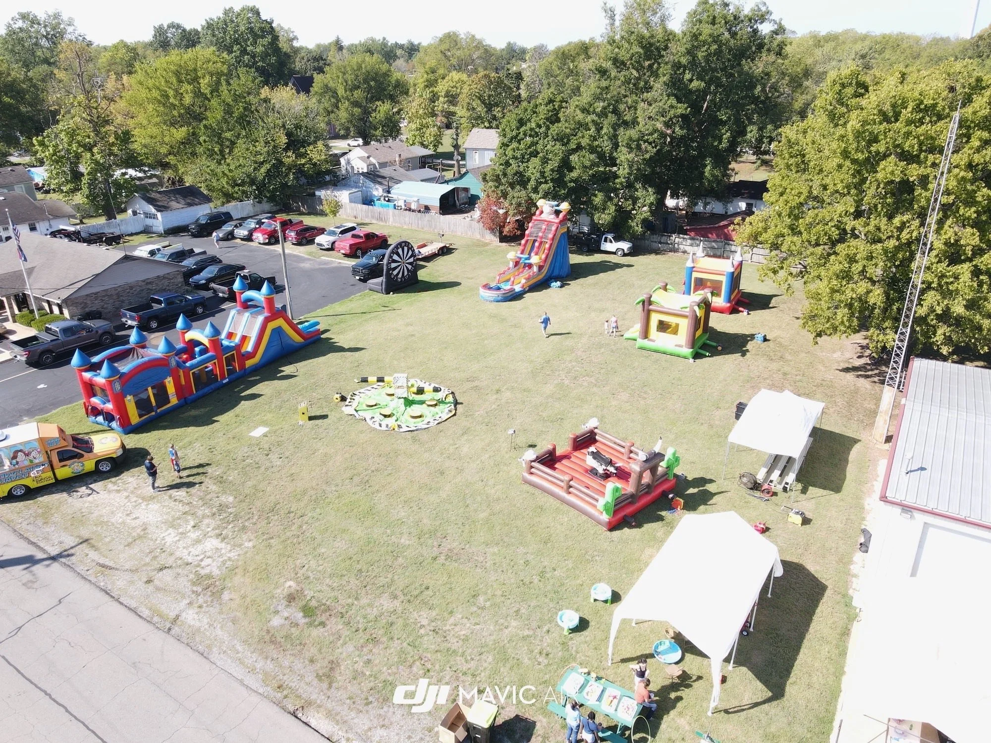 Aerial view of a children's outdoor amusement area with inflatable slides, bounce houses, a carnival game zone, and tents, surrounded by cars, houses, and trees.