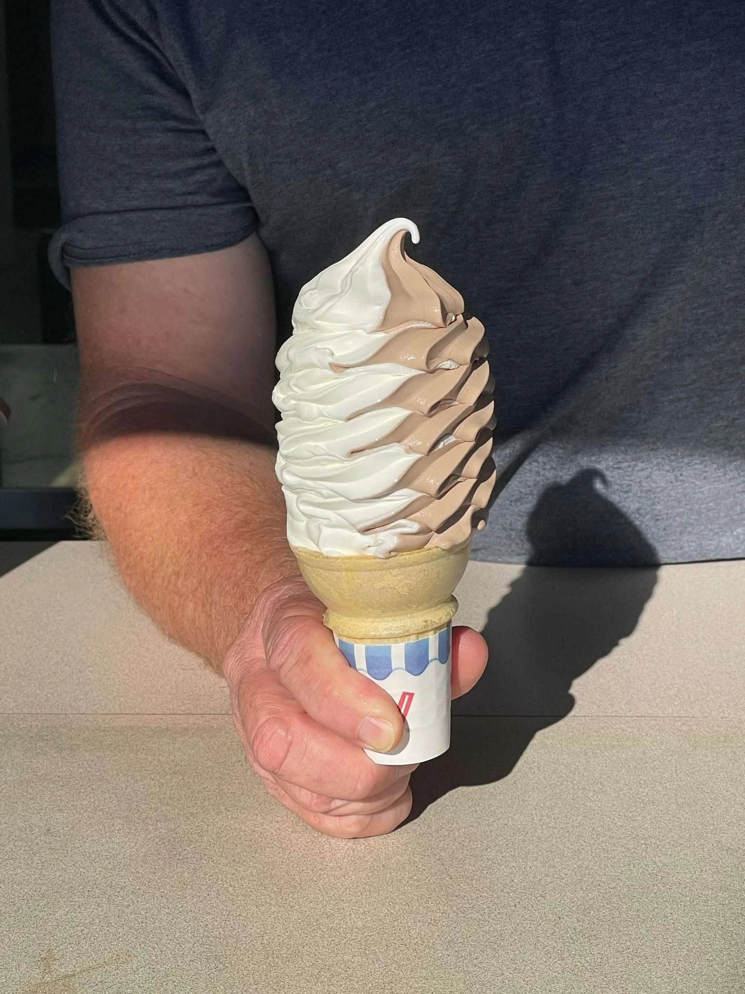 A person holding a large soft serve ice cream cone with vanilla and coffee flavored swirls, standing at a table.