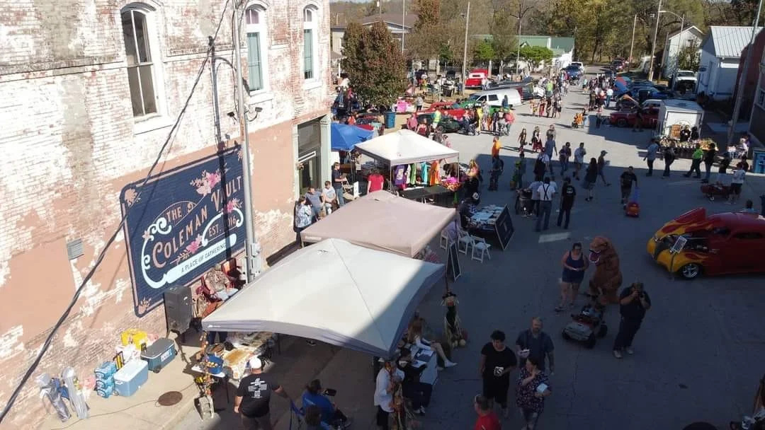 Outdoor street fair with vendor tents, people browsing, and vintage cars parked nearby.