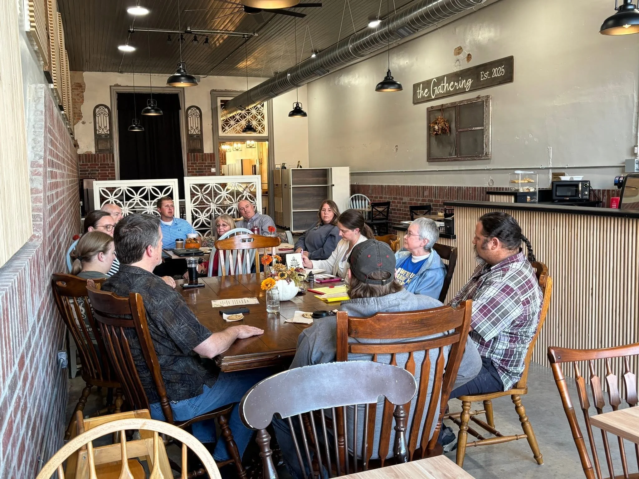 People sitting around a large wooden table in a cozy restaurant with brick walls and industrial ceiling, engaged in conversation and looking towards the front.