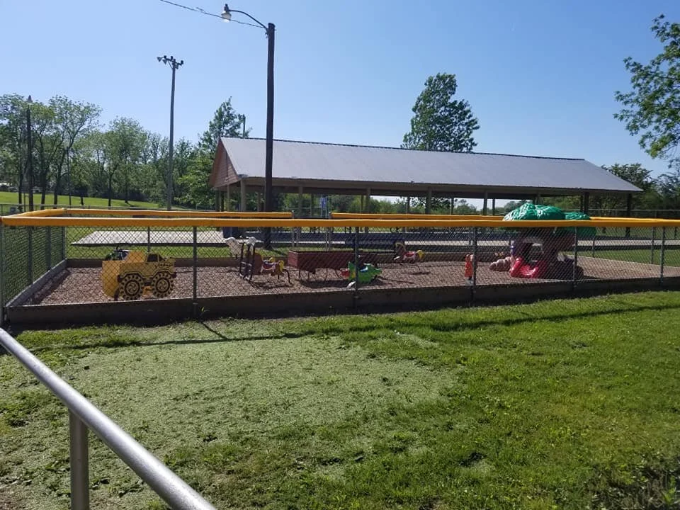 Children's playground with sandbox, small rocking horses, and a play structure with a slide, enclosed by a black chain-link fence.