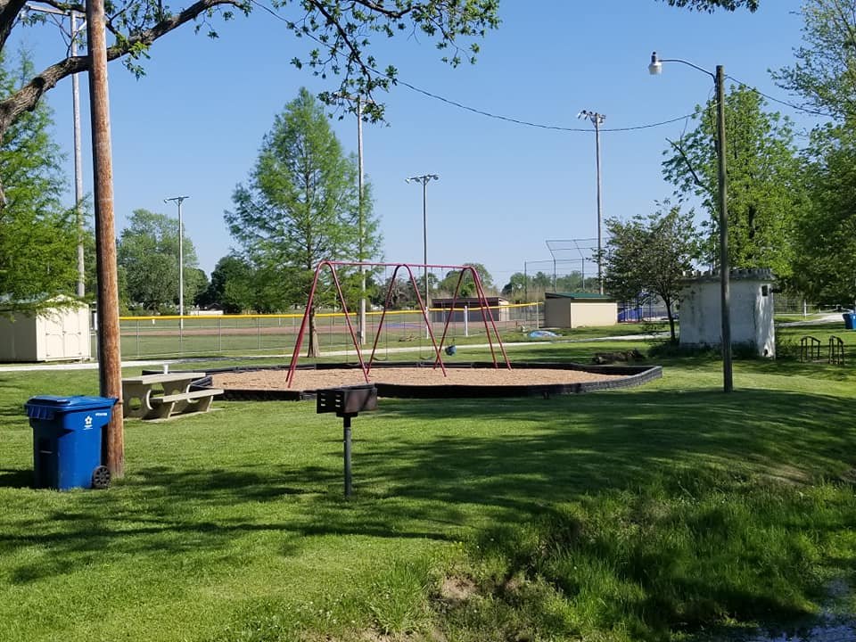 A park playground with swings on a round sand area, surrounded by green grass, trees, and sport fields in the background.