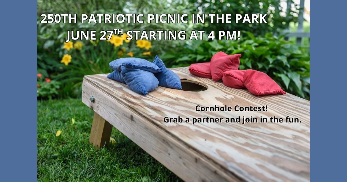 A cornhole board with blue and red bean bags on it, set outdoors on grass with flowers and greenery in the background, advertising a patriotic picnic and cornhole contest.