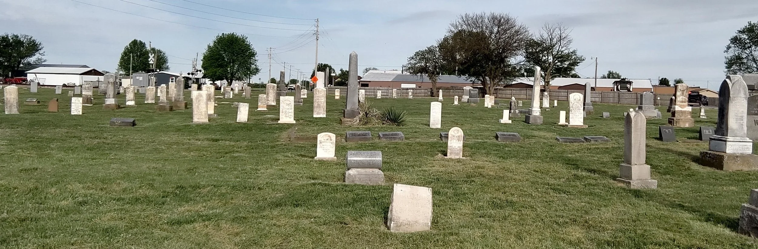 A cemetery with numerous white and gray headstones and monuments on a green grassy field, with trees and buildings in the background under a partly cloudy sky.