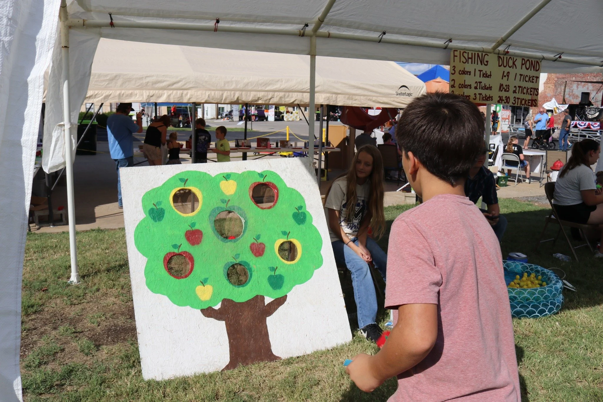 A boy playing a ring toss game at an outdoor fair with a tree painted on a board and holes as targets. There are tents and people in the background.