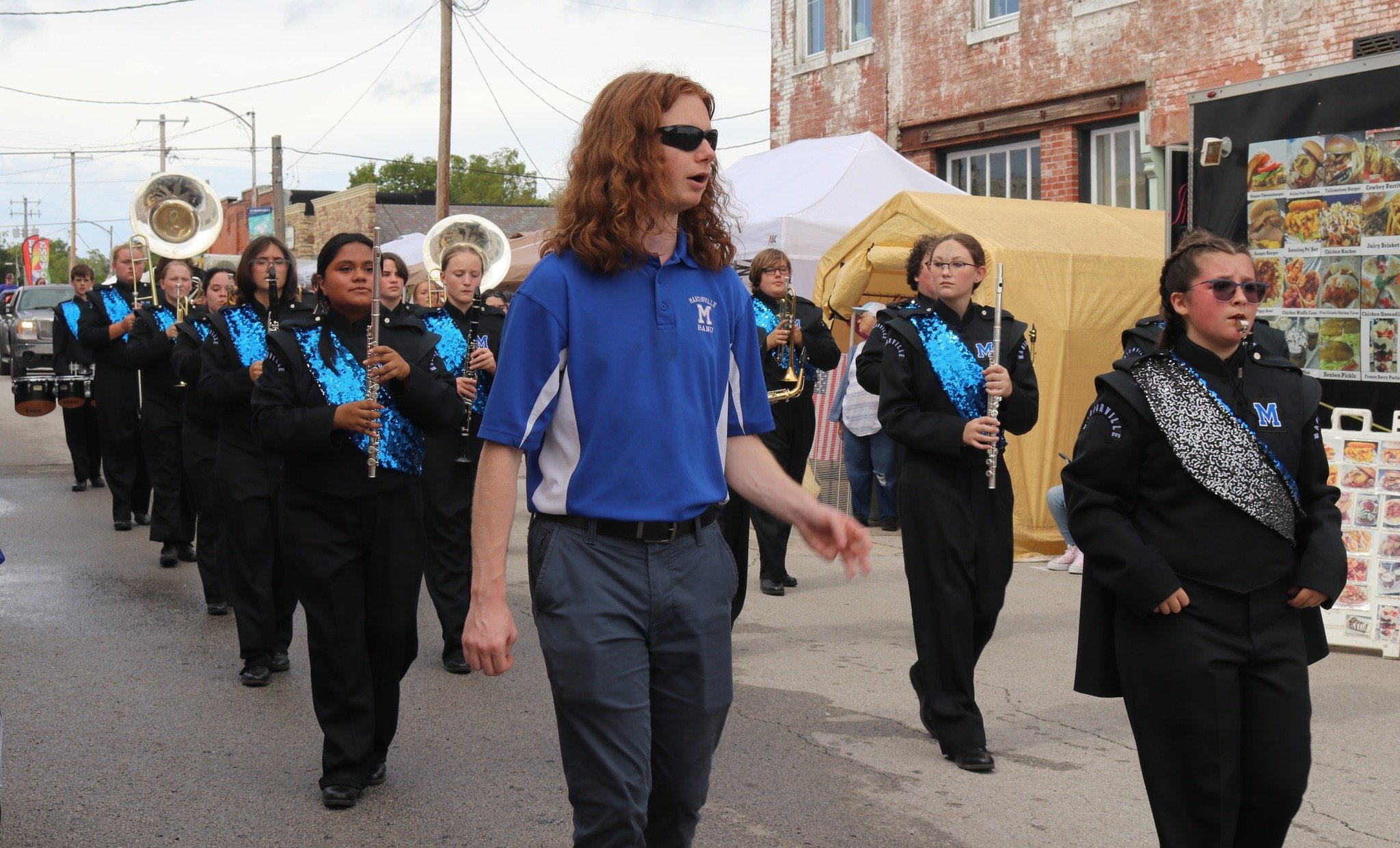 A parade featuring a marching band with young female musicians, led by a woman in sunglasses and a blue polo shirt, walking on an urban street with tents and food stands in the background.